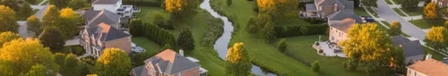 Aerial view of a quiet suburban neighborhood with tree-lined streets and homes bathed in warm golden afternoon sunlight.